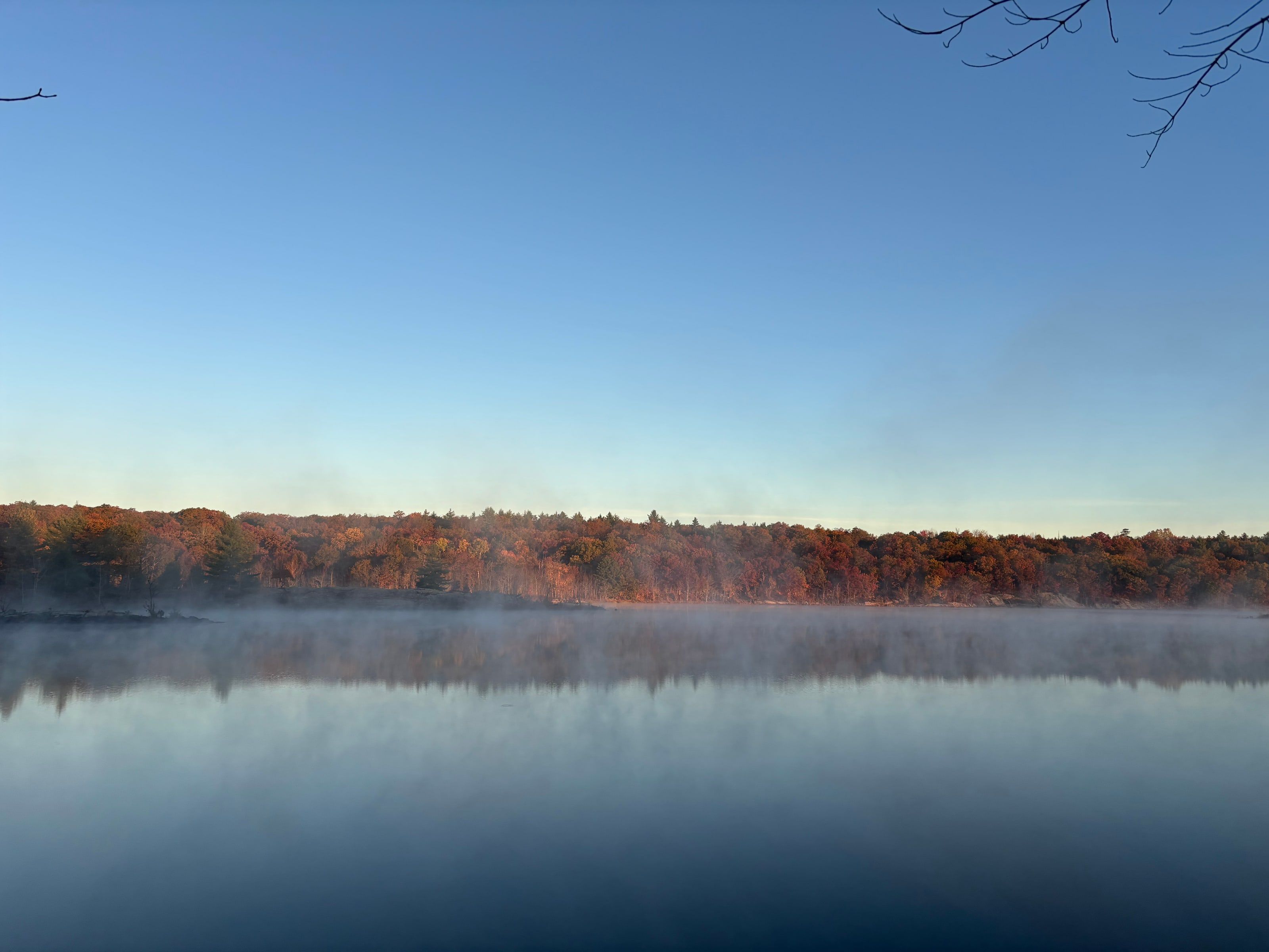 Mist rising off a still lake at sunrise, the far shore's rust and orange autumn trees mirrored in the water under a clear blue sky