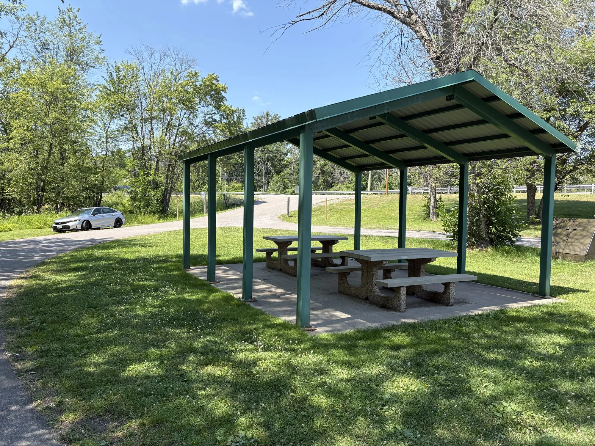 A green-roofed picnic shelter over two picnic tables on a mown lawn at the Arden Road pull-off, with the lane curving past