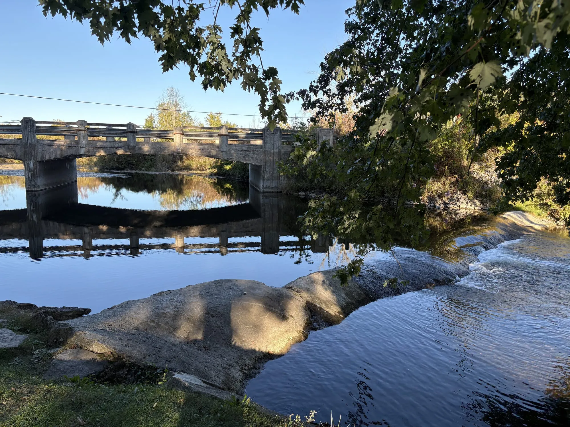 The Ouse River sliding over a low rock ledge beside an old concrete bridge in Asphodel Park, maple branches overhead