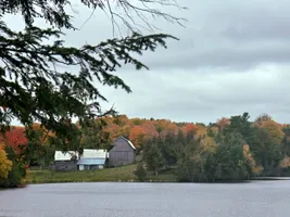 A weathered grey barn and white-roofed outbuildings across a lake under an overcast sky, with red and orange trees along the far shore and evergreen boughs in the foreground
