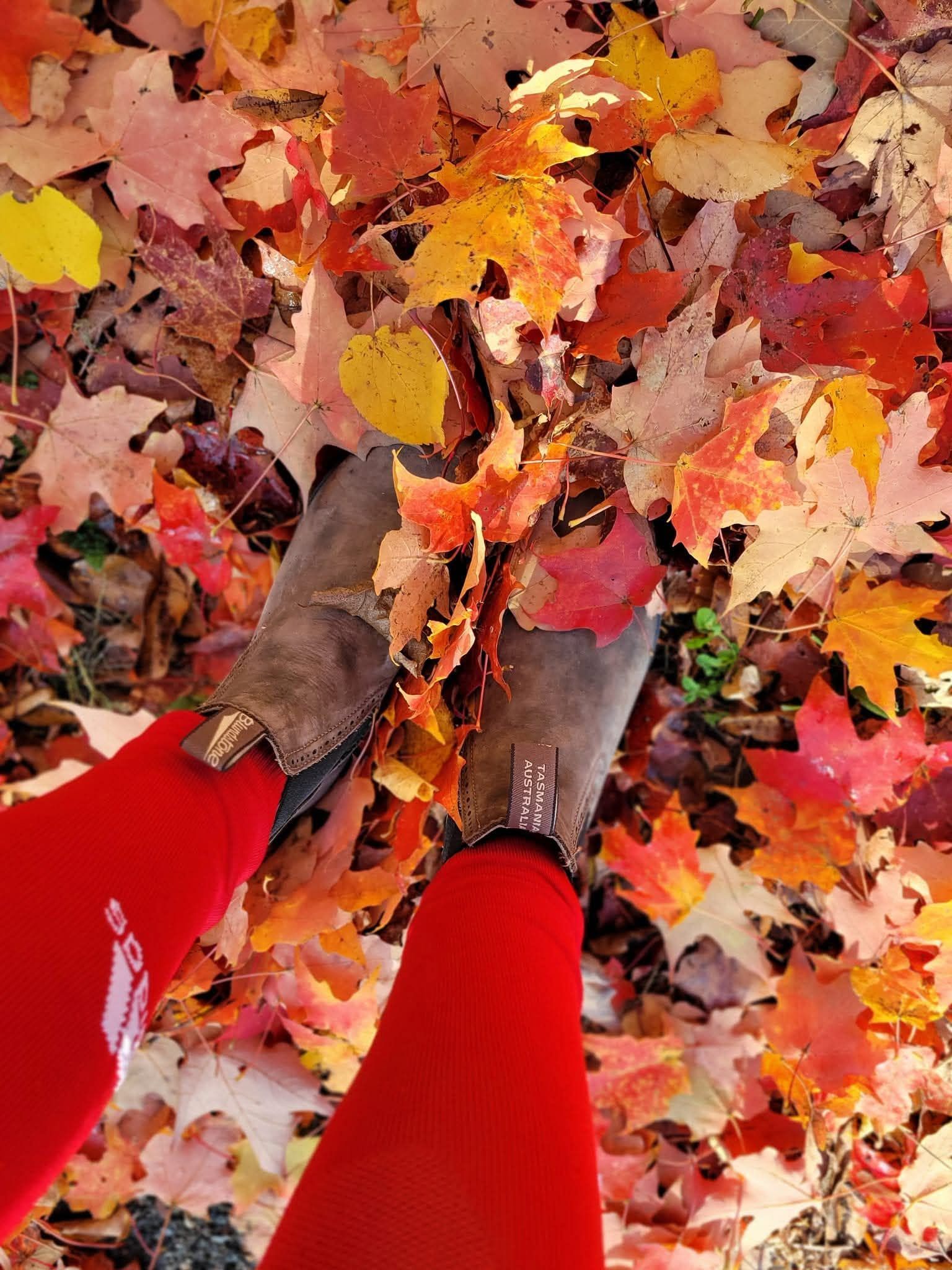 Looking down at brown boots and red leggings standing in a thick carpet of red, orange and yellow maple leaves