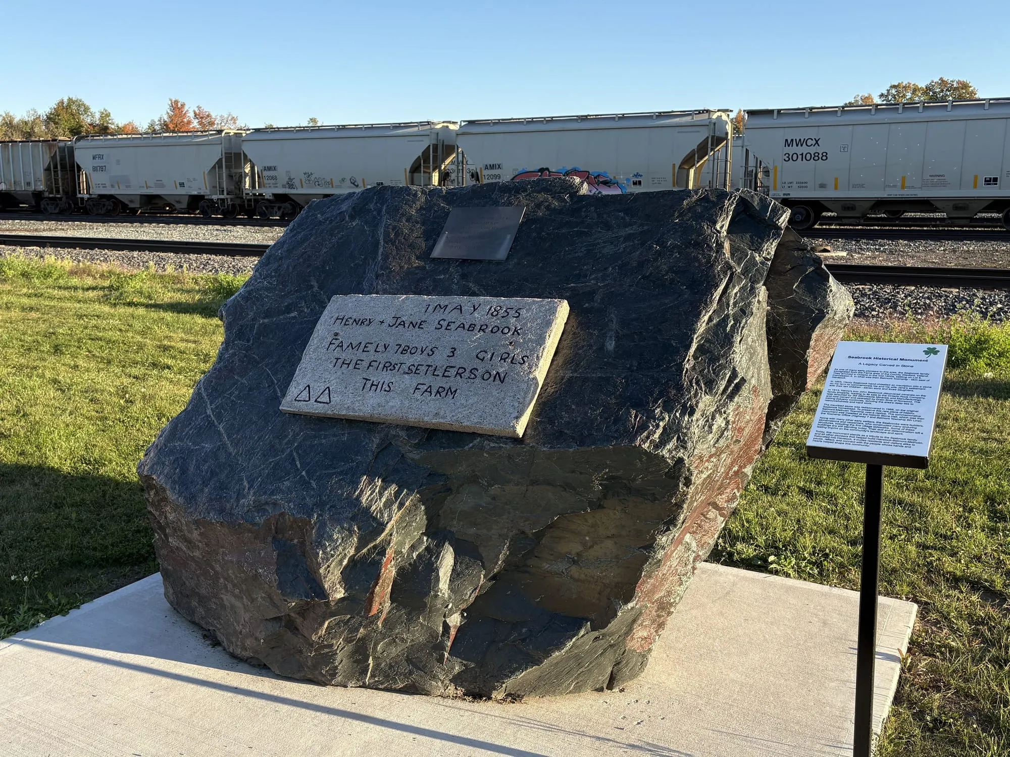 A dark boulder beside the railway in Havelock with a stone tablet reading 1 May 1855, Henry and Jane Seabrook, family 7 boys 3 girls, the first setlers on this farm, and an interpretive sign alongside