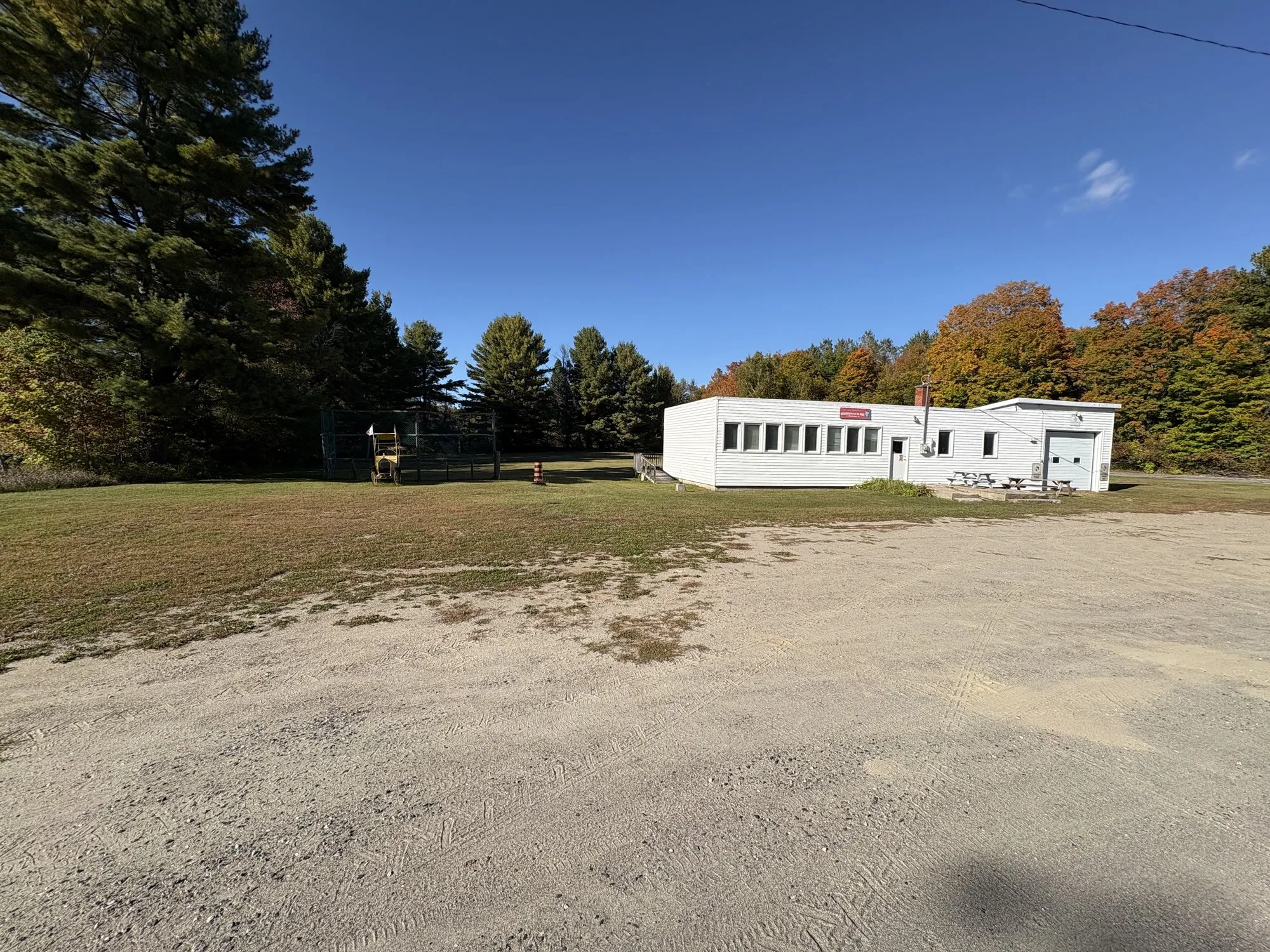 The Henderson UCW Hall, a long low white building with a row of windows, set back across a gravel lot with fall colour in the woods behind