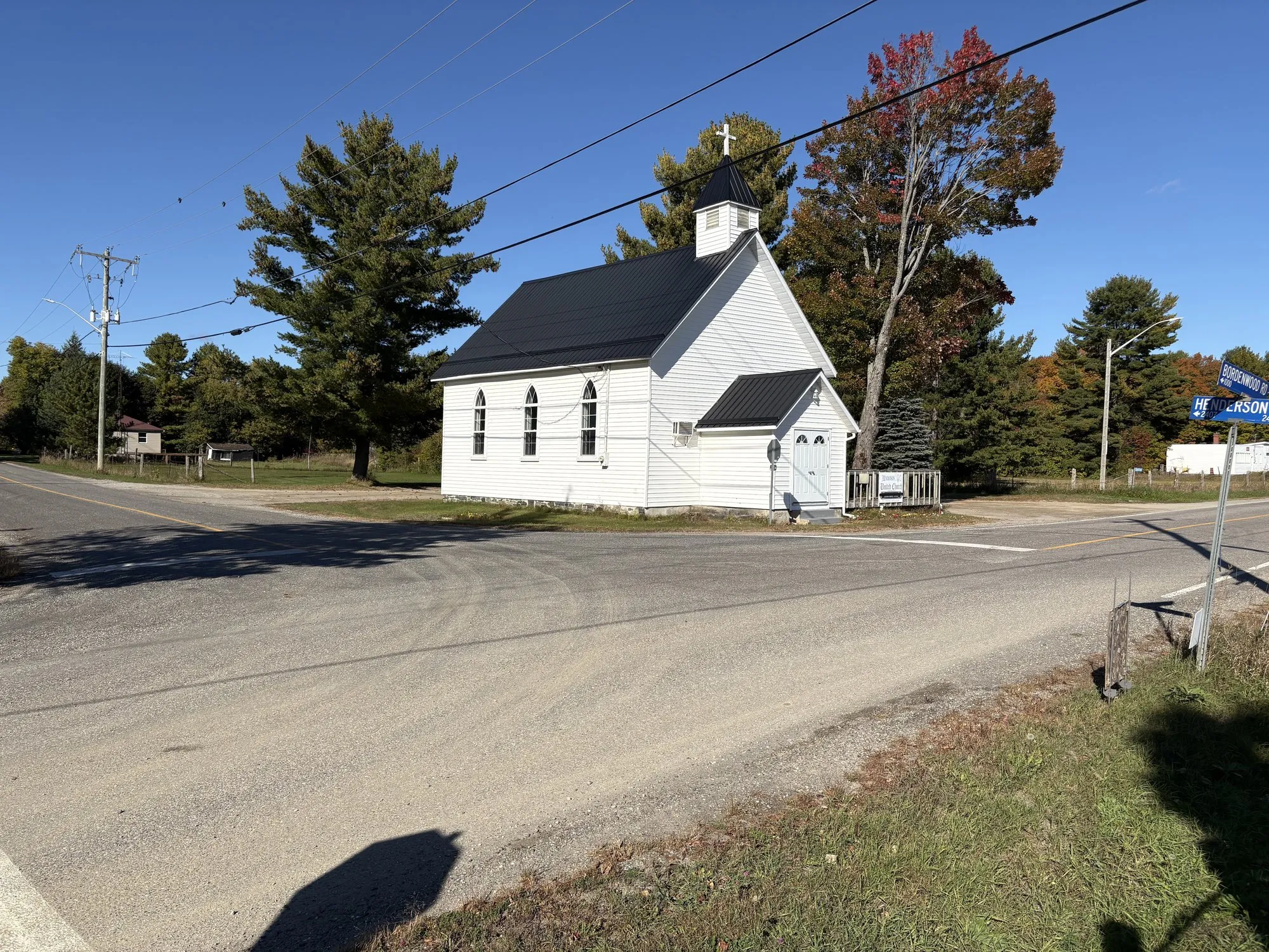 Henderson United Church, a small white clapboard church with a black roof and a cross on its belfry, at the corner of Henderson Road under a big pine