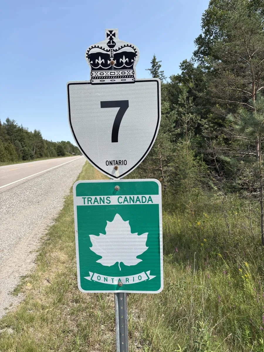 The crowned Ontario Highway 7 shield mounted above a green Trans-Canada Highway marker, on the gravel shoulder with the road running away behind it