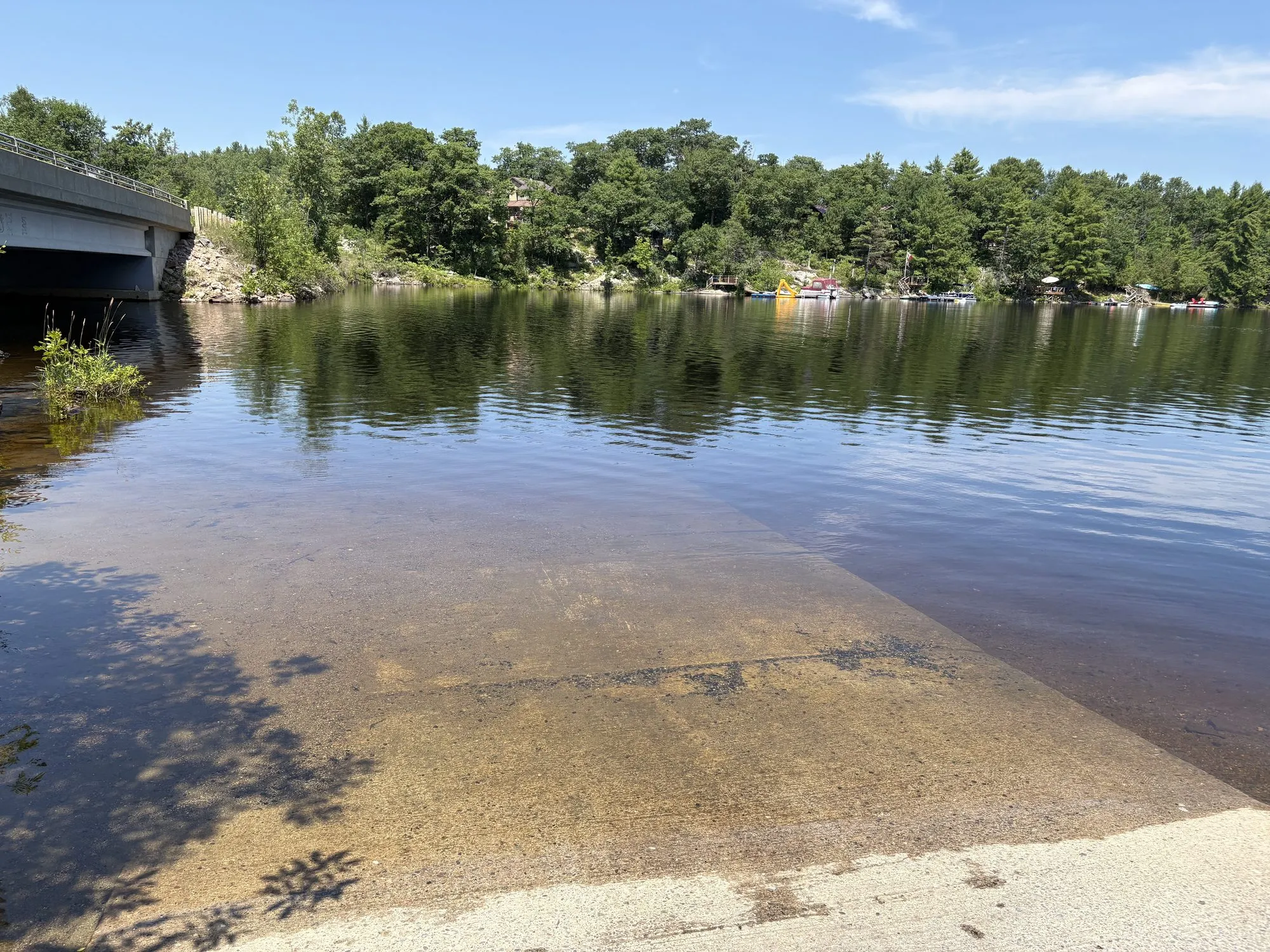 A concrete boat ramp sloping into Kennebec Lake beside the Henderson Road bridge, cottages and moored boats on the far shore