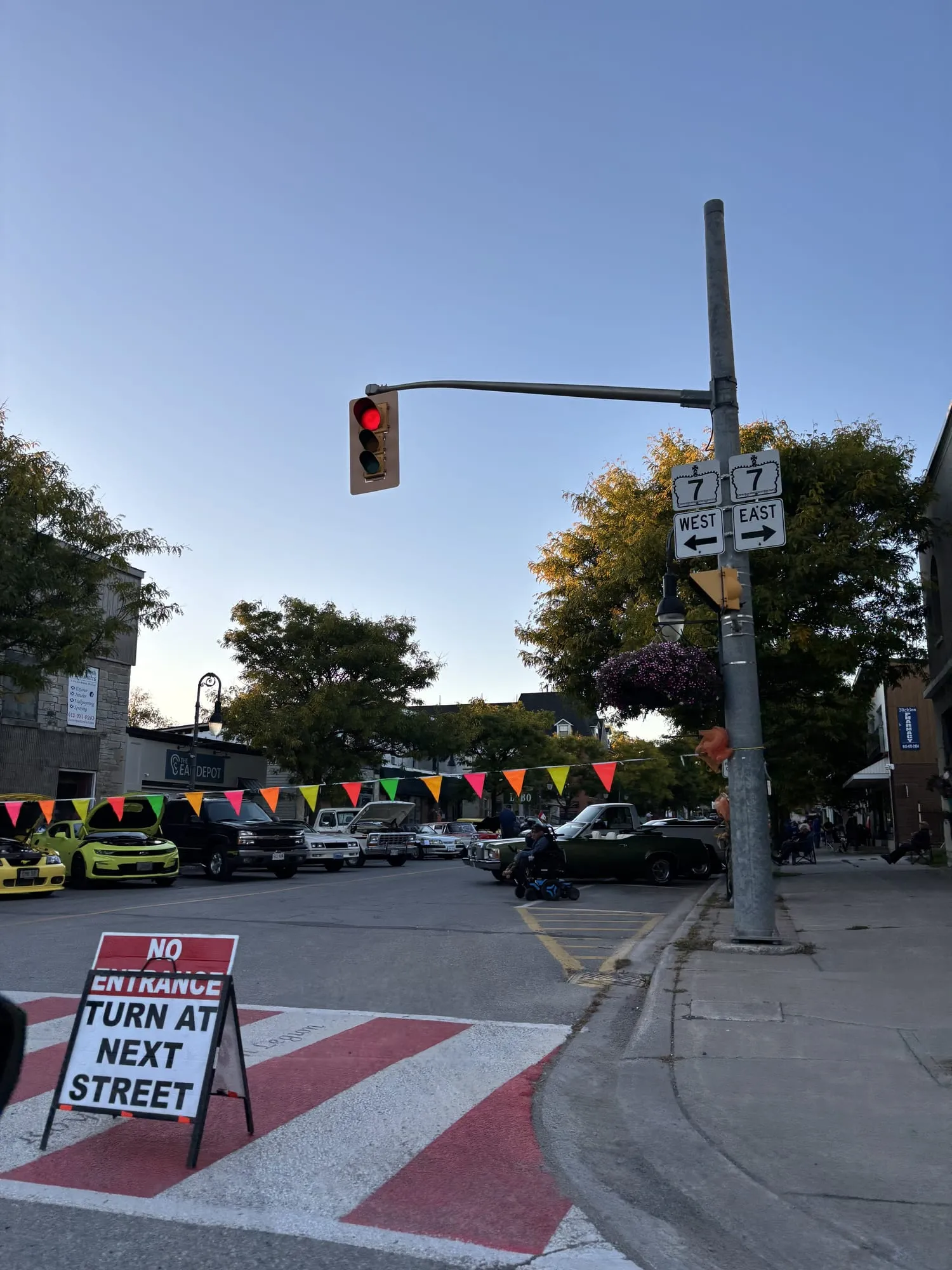 Hwy 7 East and West route signs on a traffic-light pole at the corner of Forsyth Street in Marmora, classic cars parked under strings of pennants on the street behind