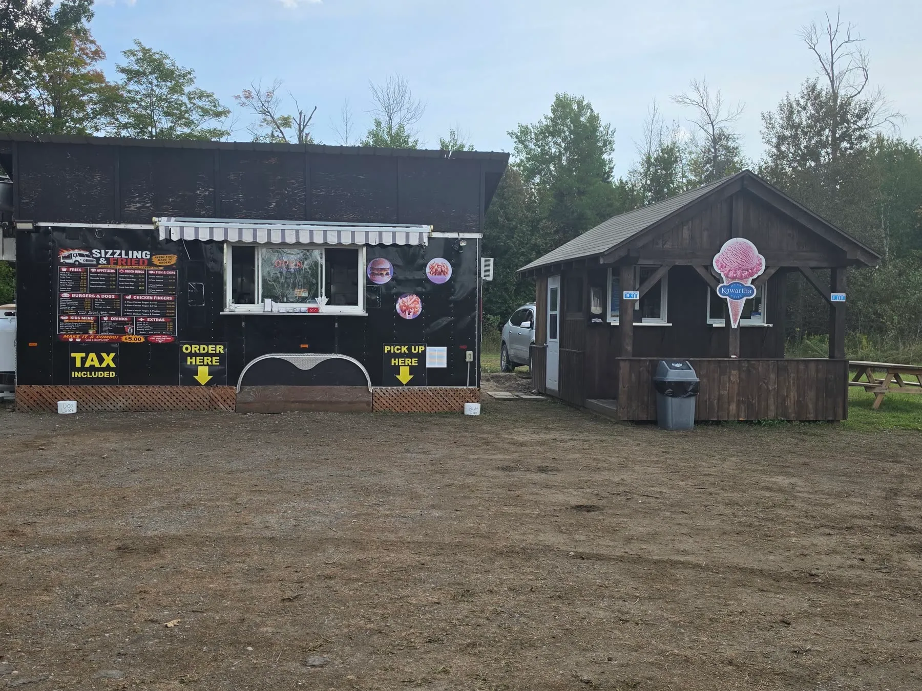The black Sizzling & Fried chip truck with its menu board and awning, beside a small wooden ice cream cabin with a Kawartha Dairy cone sign, on a gravel lot