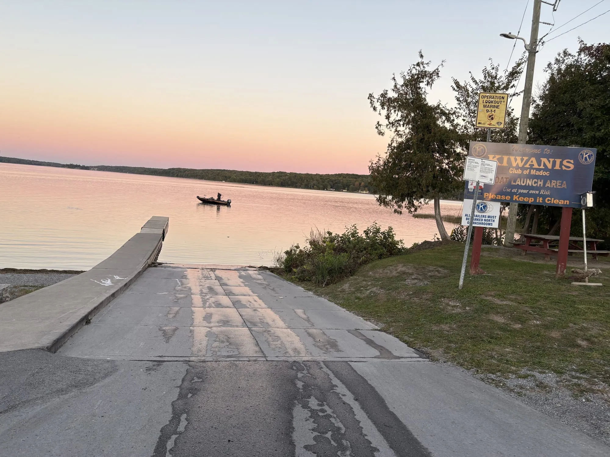 The concrete ramp at the Moira Lake launch running into a pink evening lake, a fishing boat offshore and the Kiwanis Club of Madoc boat launch sign on the grass