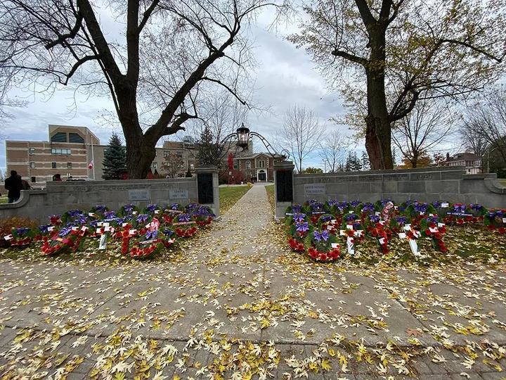 Wreaths of poppies laid along both wings of the Perth cenotaph, its stone walls marked 1914, 1939 to 1945, Korea, Afghanistan and United Nations Peacekeeping, with a path through the gates to the Great War Memorial Hospital behind and fallen leaves across the walk.