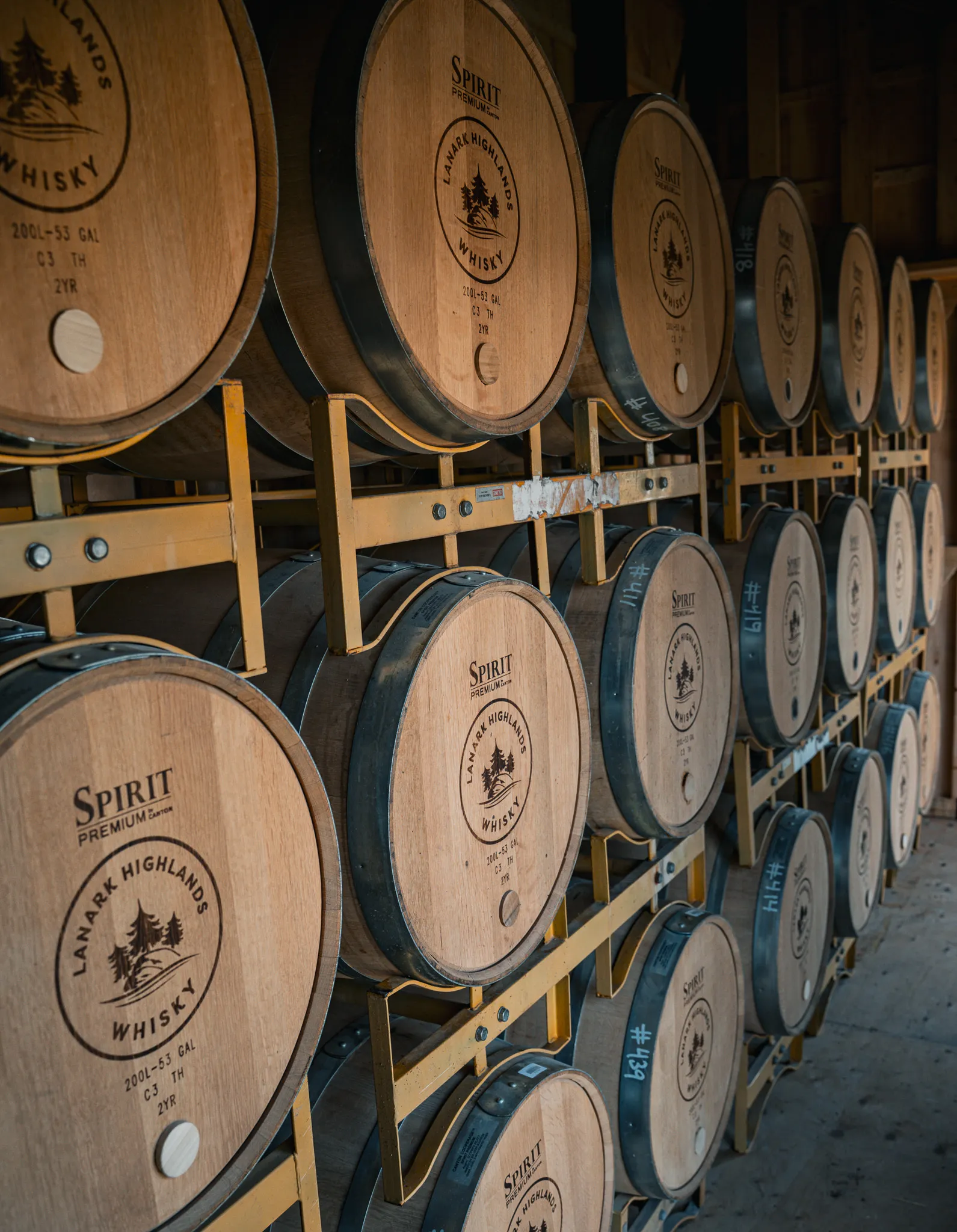 Rows of whisky barrels on yellow steel racks in the Top Shelf Distillers barrel room, the heads branded Lanark Highlands Whisky