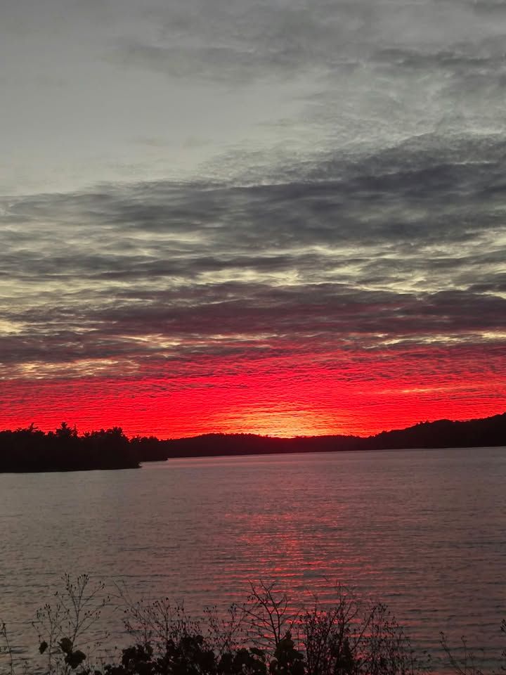 A band of red sunset light under heavy grey cloud, reflected in a lake below a dark wooded shore