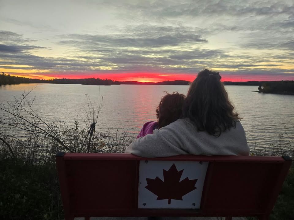 A woman and a child seen from behind on a red bench with a white maple leaf, looking out over a lake at a red sunset