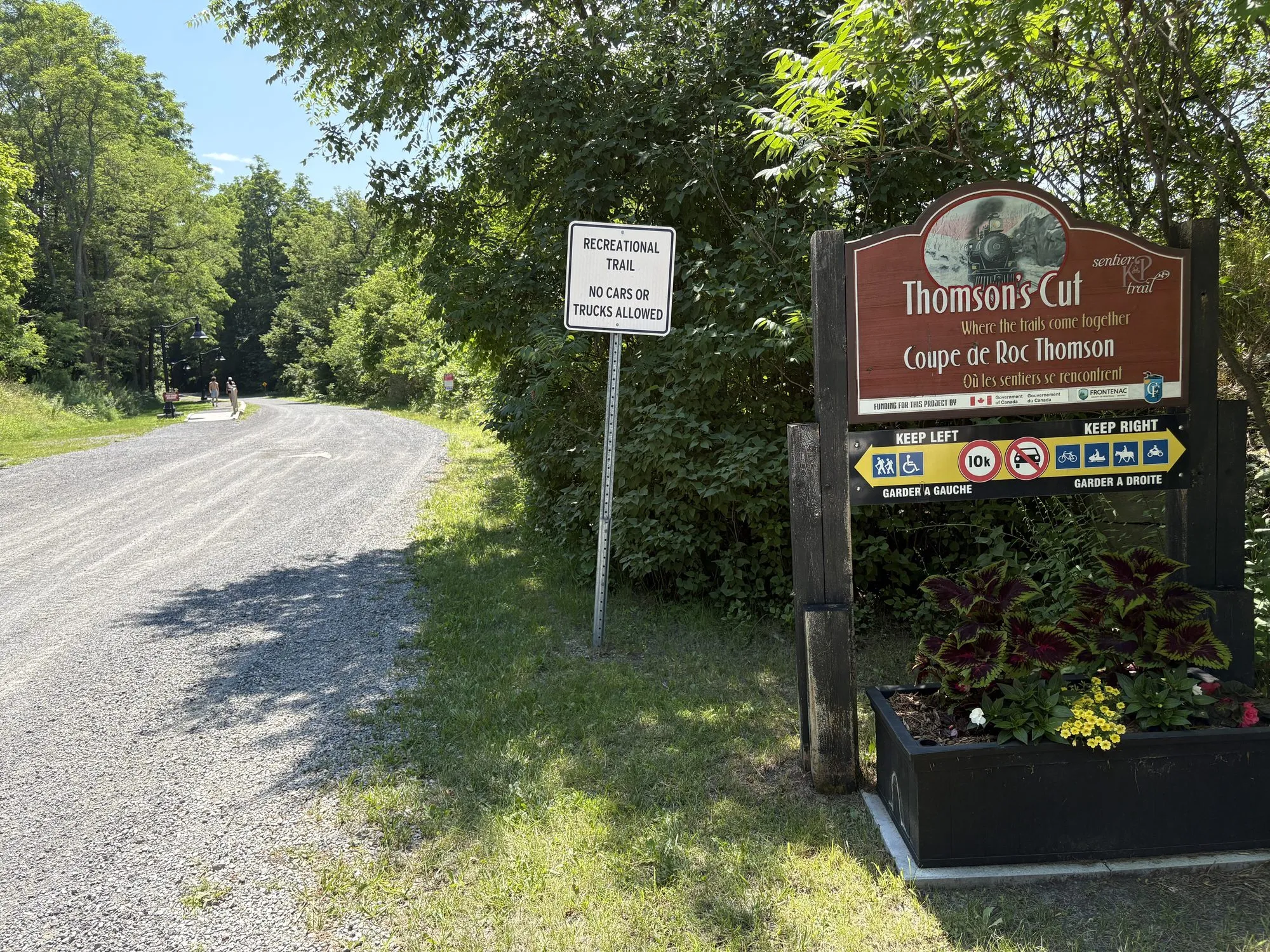 The trailhead sign at Thomson's Cut where the K&P and Trans Canada trails meet, stone-dust trail curving away under the trees