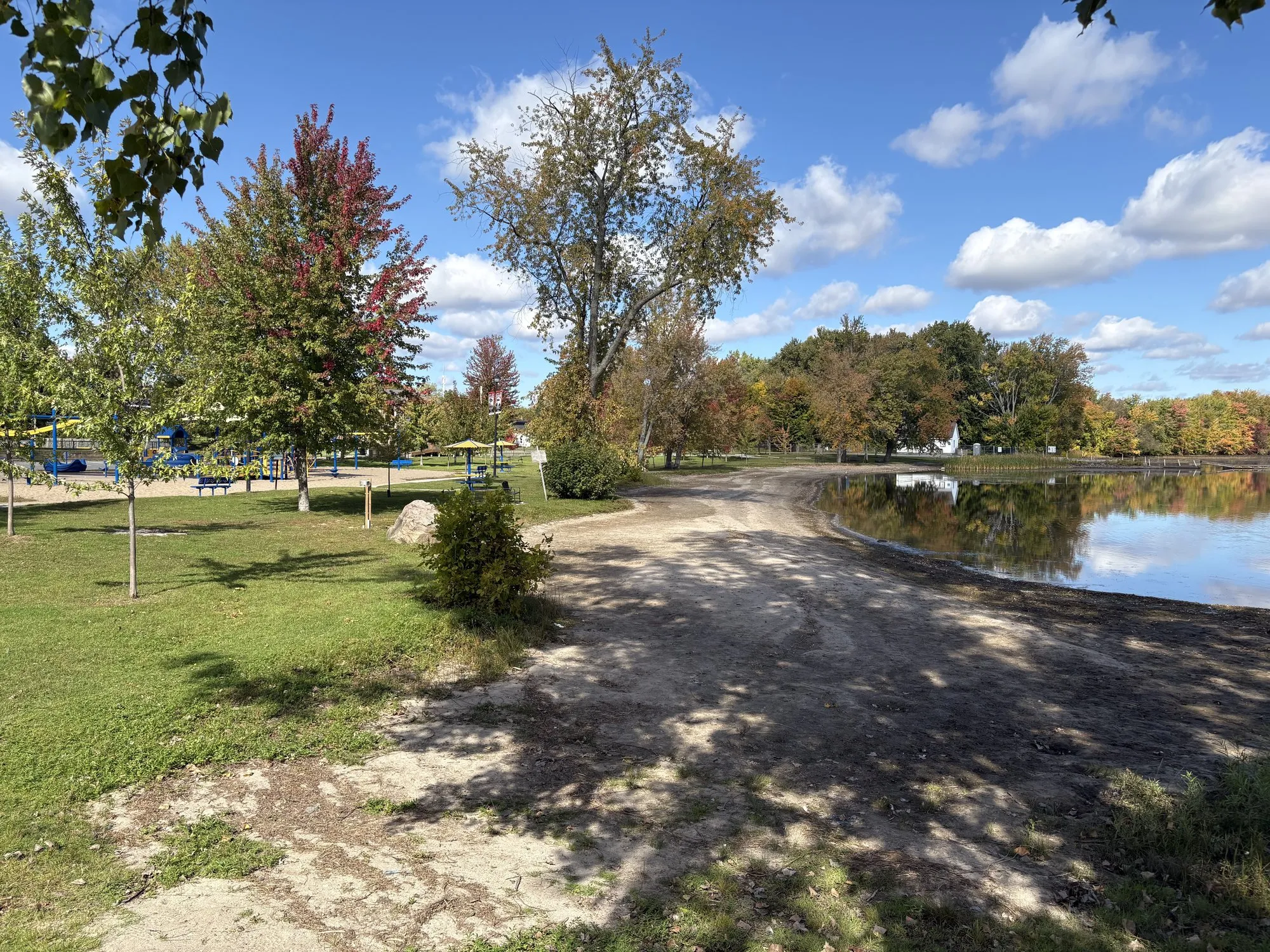 A sandy path curving down to Stoco Lake at Tweed Memorial Park, a red maple and the playground on the left and fall colour along the far shore